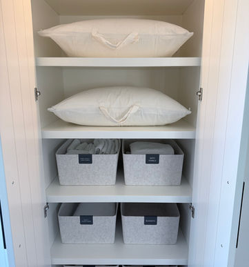 A guest room linen cabinet with pillows in canvas storage bags on the top two shelves, and linens in felt baskets on the bottom three shelves.