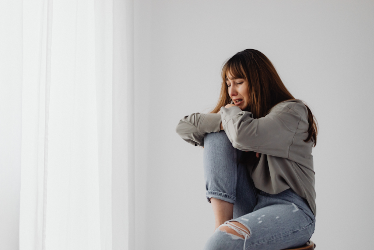 Woman in gray sweater and ripped jeans sitting, looking sad, in a bright, minimalist room with white curtains.