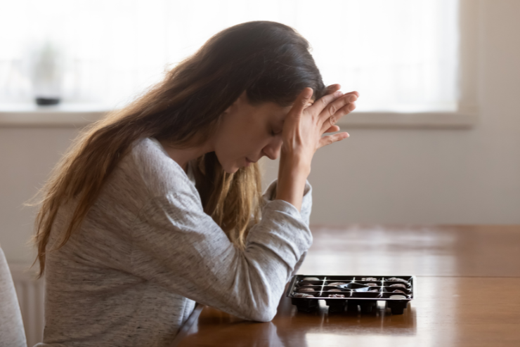 Woman in a gray sweater sits at a table, resting her head in her hands in contemplation. An open chocolate box is nearby. Soft daylight fills the room.