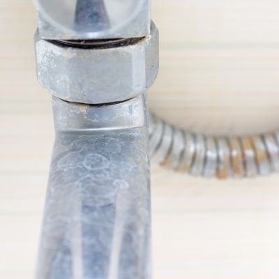 Close-up of a tarnished metal pipe and connector against a light wood background. The pipe shows signs of wear and mineral deposits.