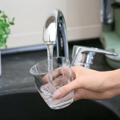 Hand holding a glass under a shiny chrome faucet with water flowing. Black countertop, green plant in the background, kitchen sink setting.
