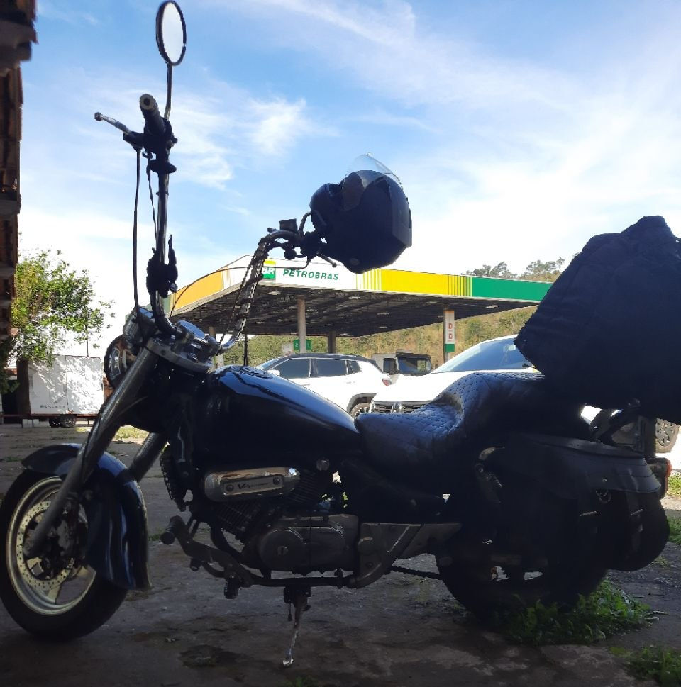 A foto de Marruá, uma moto custom preta com guidão seca suvaco de frente a um posto Petrobrás