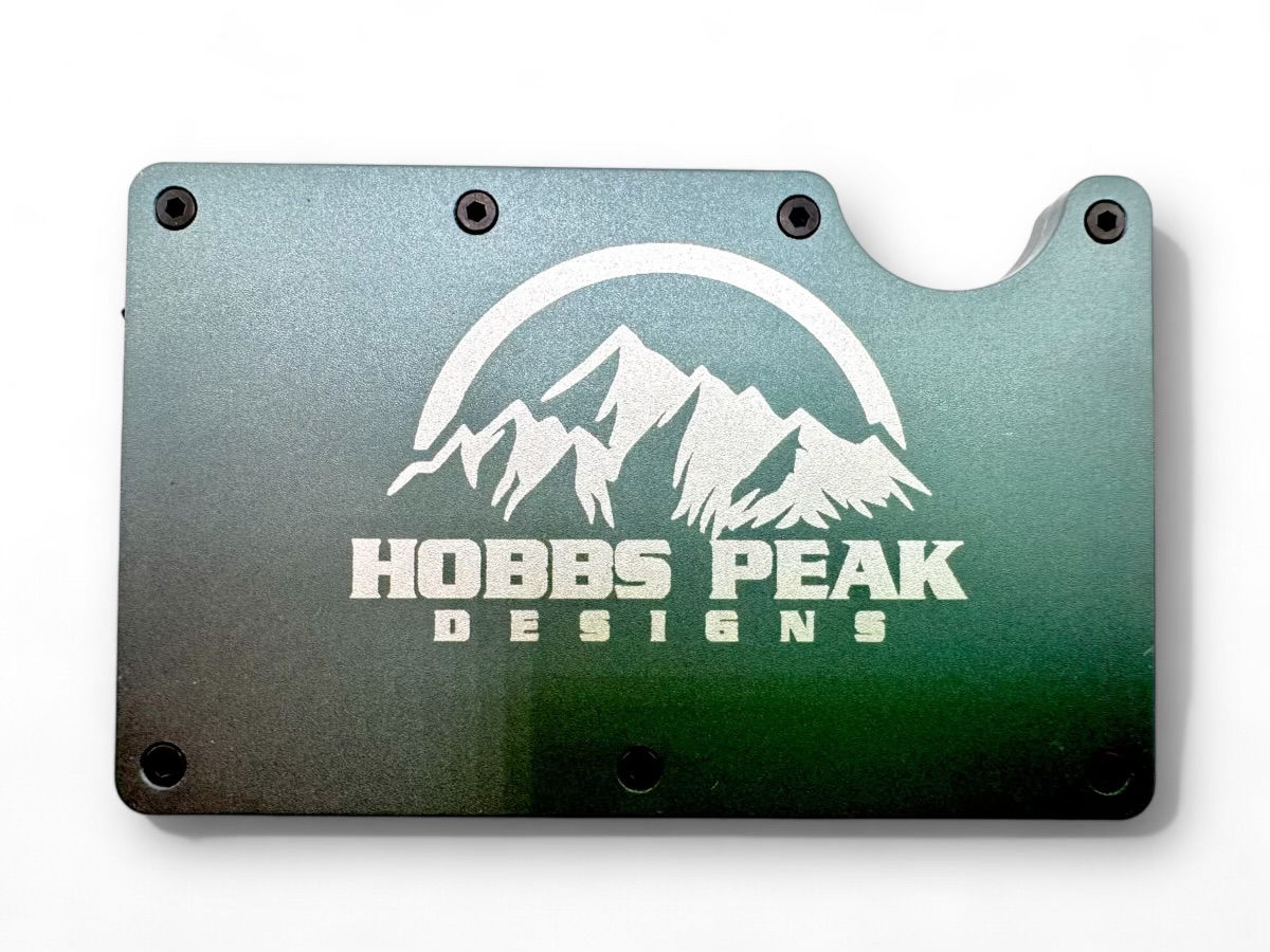 "The image shows a metallic object with a gradient color scheme transitioning from dark green at the bottom to a lighter green at the top. The object has a logo and text engraved on it. The logo features a stylized mountain range with a semi-circular arc above it. Below the logo, the text reads 'HOBBS PEAK DESIGNS.' The object has six holes, presumably for screws or bolts, with one of the corners having a cut-out section. This image is interesting as it appears to be a custom-designed metal plate, possibly for branding or decorative purposes.