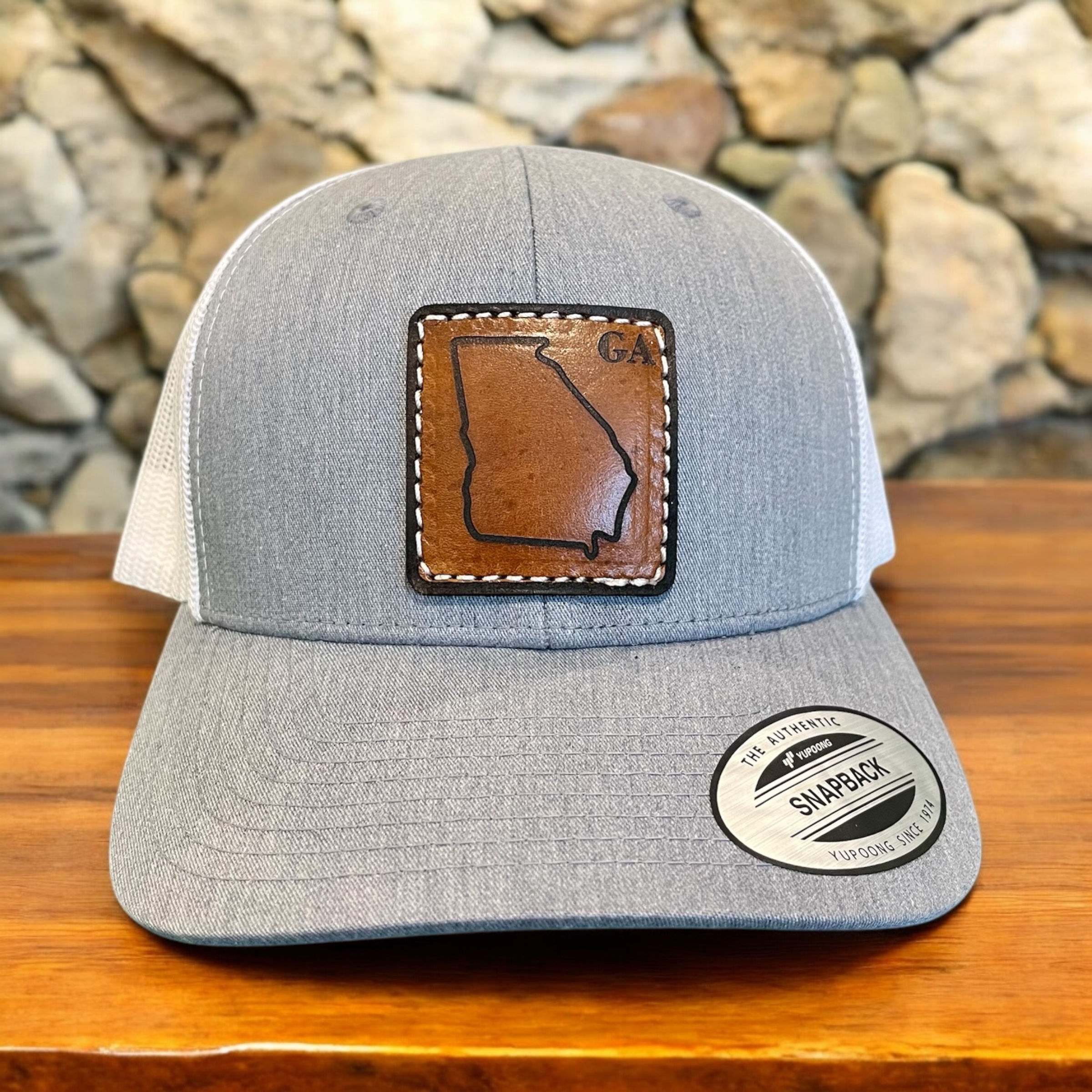 The image shows a gray and white baseball cap with a leather patch on the front. The leather patch features an outline of the state of Georgia with the letters "GA" in the top right corner. The cap has a sticker on the brim that reads "THE AUTHENTIC SNAPBACK YUPOONG CLASSICS." The background consists of a stone wall, and the cap is placed on a wooden surface