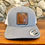 Thumbnail: The image shows a gray and white baseball cap with a leather patch on the front. The leather patch features an outline of the state of Georgia with the letters "GA" in the top right corner. The cap has a sticker on the brim that reads "THE AUTHENTIC SNAPBACK YUPOONG CLASSICS." The background consists of a stone wall, and the cap is placed on a wooden surface