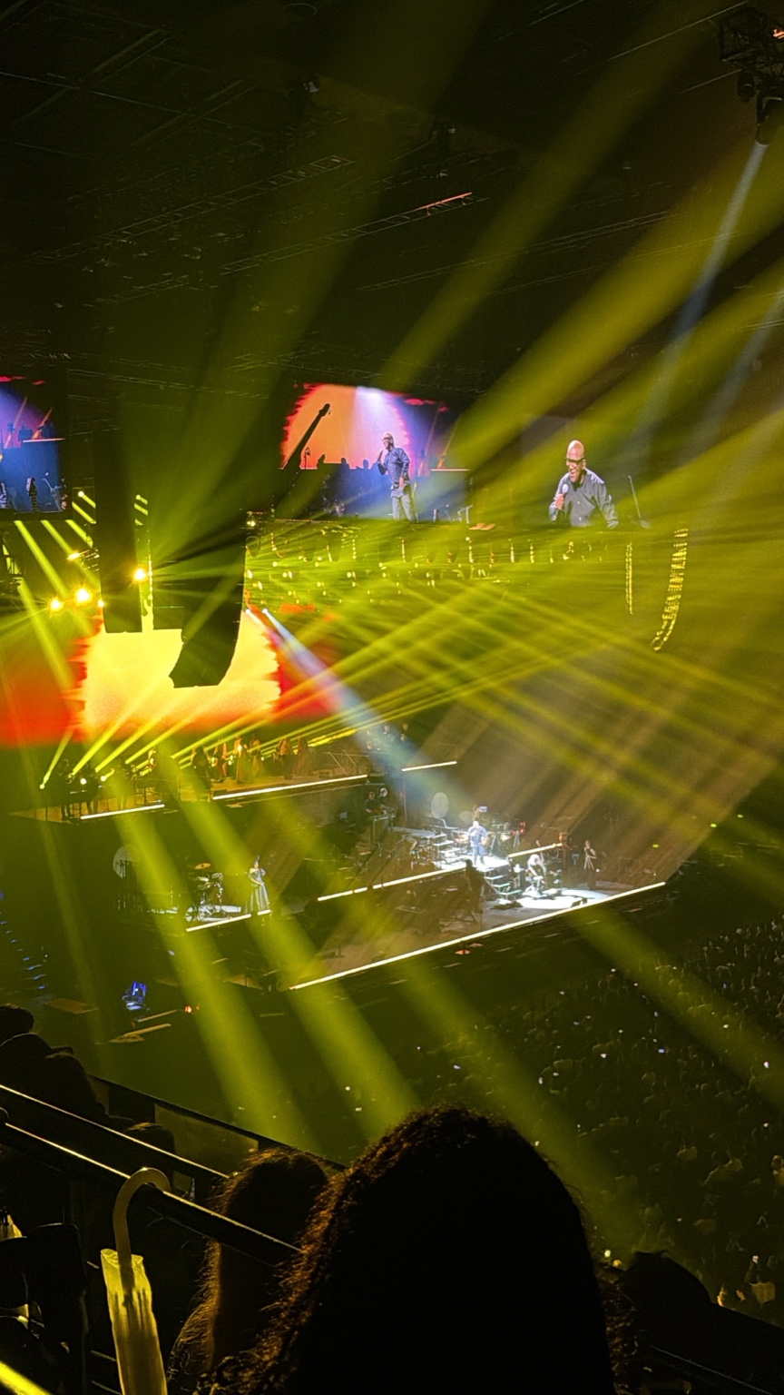 A concert stage beneath sweeping yellow light beams, with large screens above the musicians.