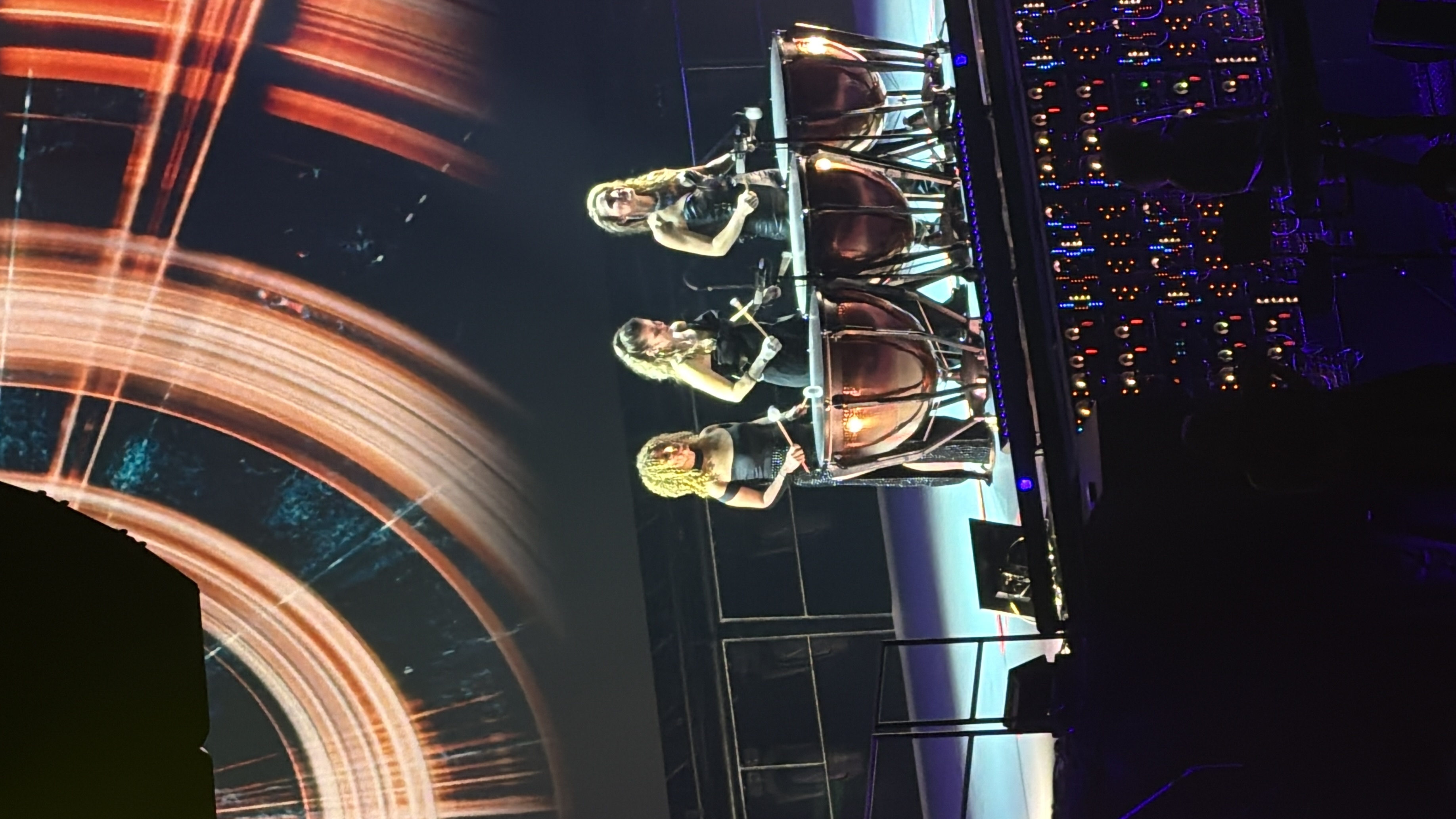 Three percussionists hold mallets beside large timpani beneath a concert screen.