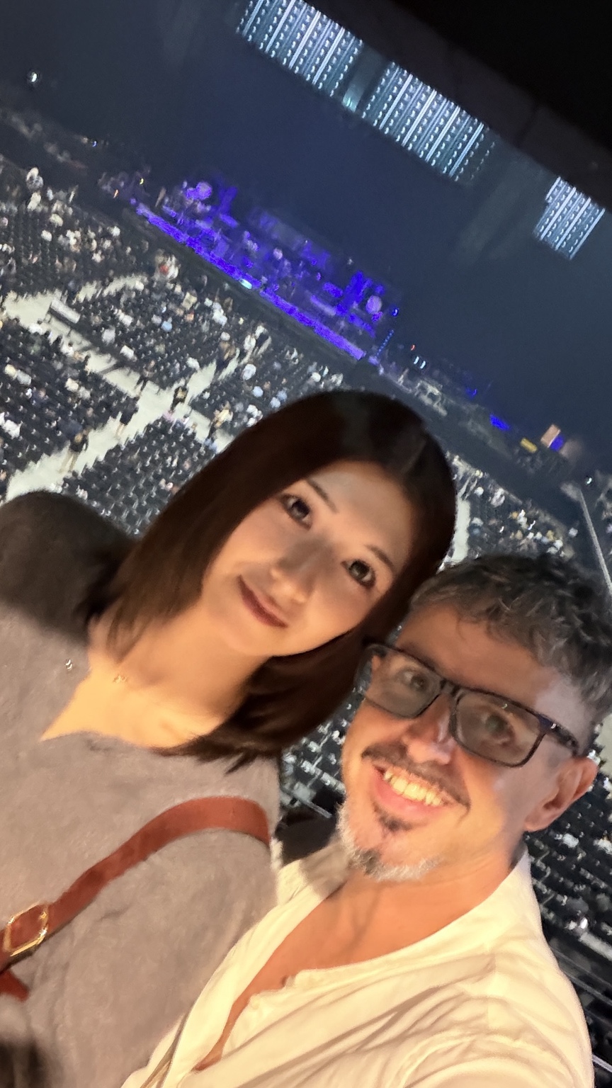 Two people smile for a close-up selfie from the arena stands, with audience seats and a stage behind them.