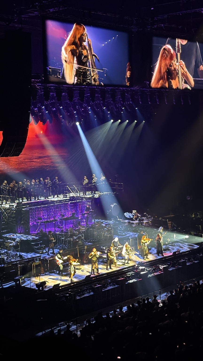 Musicians perform on a concert stage while a bowed string player is shown on large overhead screens.