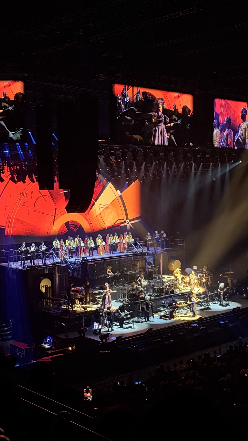 A band and choir perform on multiple stage levels beneath red and orange concert screens.