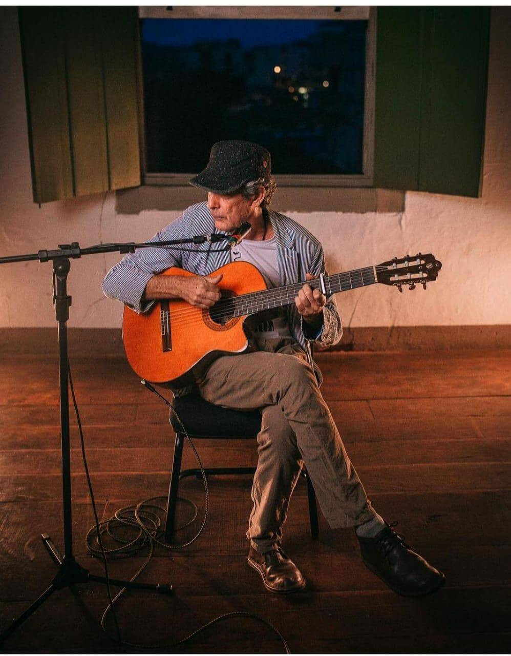 Tuca está sendo em uma cadeira, tocando violão e cantando. Ele usa uma blusa social aberta, calça marrom, sapatos sociais e uma boina.
