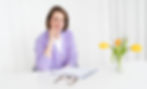 Freundlich lächelnde Frau Sillem in lila Strickjacke am weißen Tisch, in einem weißen Raum. Auf dem Tisch befindet sich ein Vase mit drei gelben Tulpen und ein Notizbrett und die Brille. Mit aufmerksamen freundlichen Blick hört Frau Katrin Sillem zu.