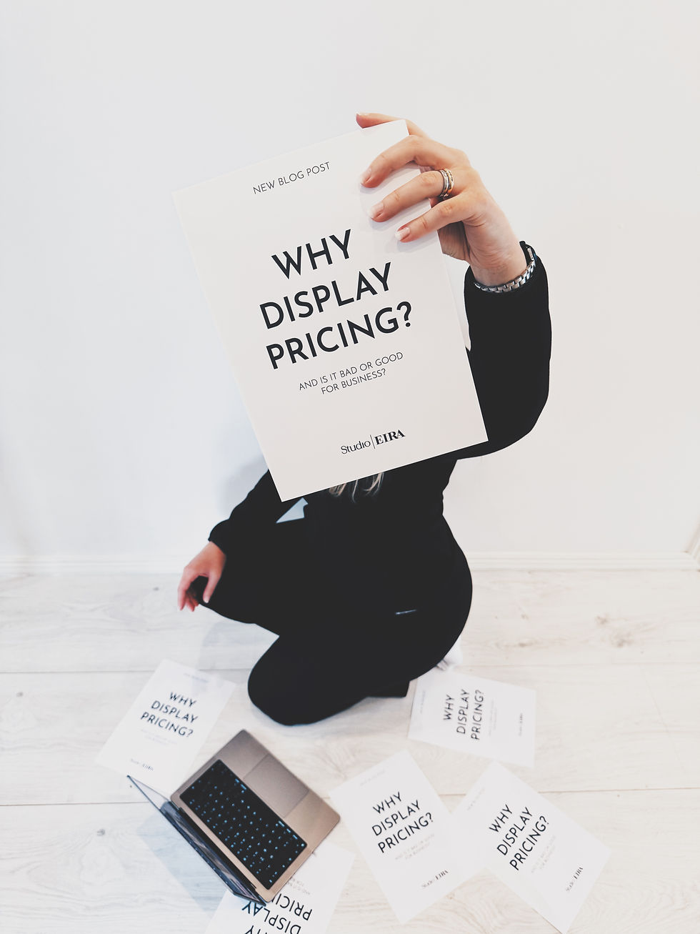Person sitter på golvet i svart outfit med laptop framför sig och håller upp ett papper med texten 'Why Display Pricing?'. Runtom ligger fler papper med samma text, mot en ljus bakgrund.