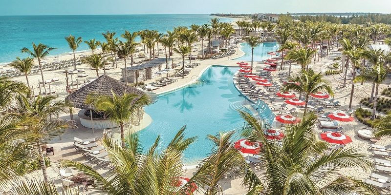 A tropical resort with a winding pool, red umbrellas, palm trees, lounge chairs, and turquoise ocean under a clear sky - bimini beach from Resorts World Bimini
