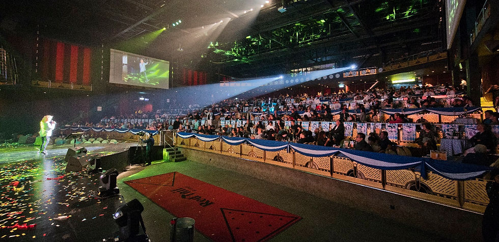 Performer on a spotlighted stage in a packed auditorium, with confetti on the floor and a large screen showing the event.