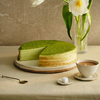 A whole Lady M Green Tea Mille Crêpes cake topped with fine matcha powder, served alongside a warm cup of tea.