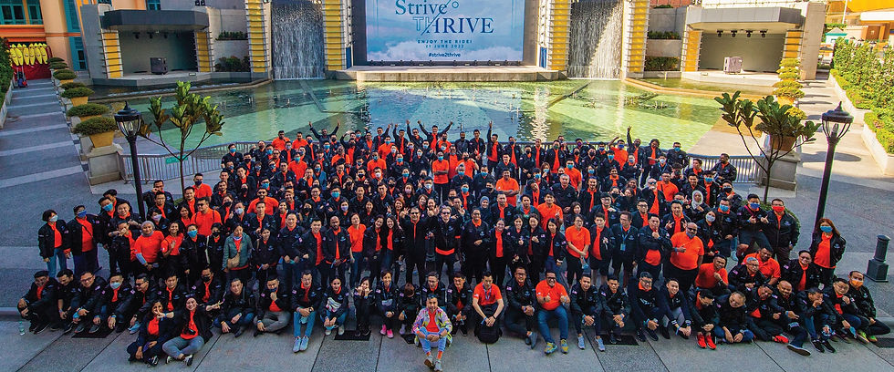 Large group in orange and black posing by a fountain at an outdoor venue, with a Strive to THRIVE banner behind them.