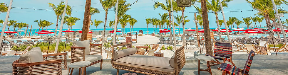 Beachside lounge with wicker furniture, palm trees, and red umbrellas. Clear blue sky and ocean in the background, creating a tropical vibe.