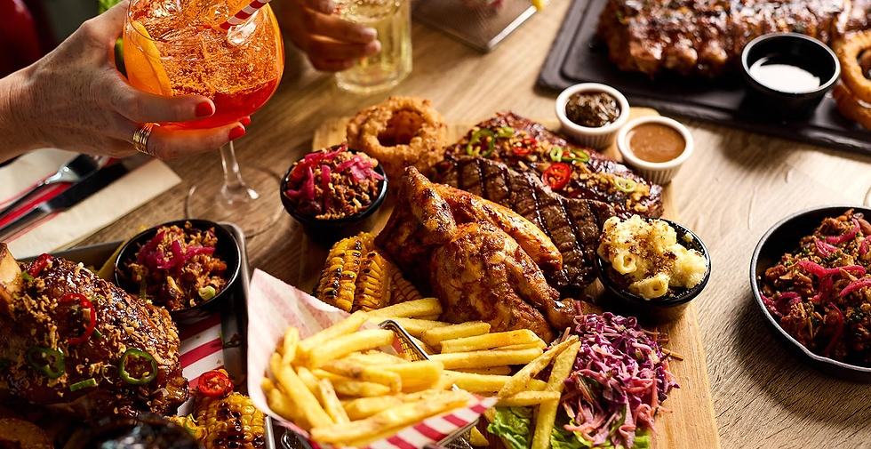 Hand holding orange cocktail over a table piled with steak, roasted chicken, fries, corn, sides, and sauces.