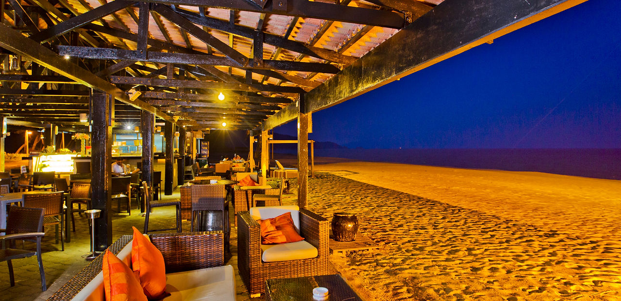 The cozy, warmly lit wooden deck of Oasis Beach Bistro overlooking the vast, dark expanse of the beach at night in Resorts World Kijal.