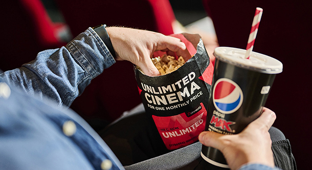 Person in a movie theater holds a popcorn bag and Pepsi cup with straw; bag reads UNLIMITED CINEMA - CINEMAX, relaxed vibe.
