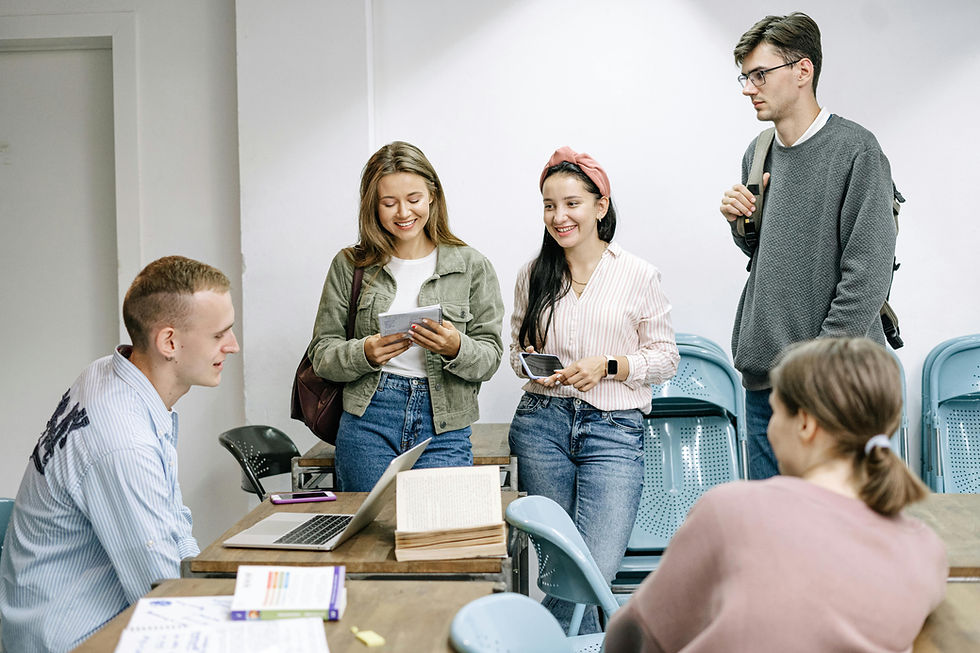 Un grupo de estudiantes en un aula, conversando y sonriendo mientras sostienen cuadernos y dispositivos electrónicos. En la mesa hay un portátil abierto y materiales de estudio. Representa un ambiente académico colaborativo.