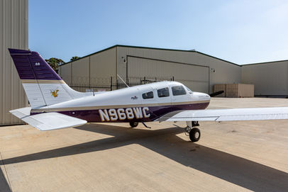 A white and purple Piper Archer III aircraft, N968WC, parked on an airfield next to hangars.