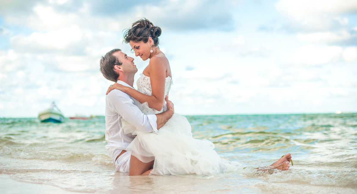 Newlywed couple during a Trash the Dress post-wedding photoshoot on Bávaro Beach in Punta Cana, with the bride sitting on the groom in the sand.