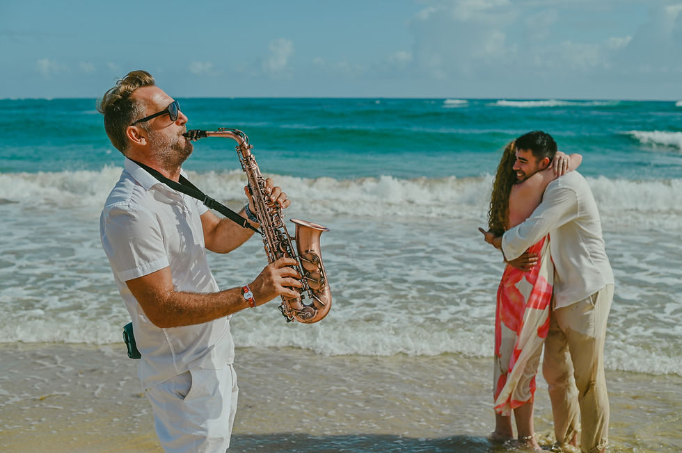 Pascal Aubrion from Punta Cana Live Music performing live saxophone music on Uvero Alto Beach in Punta Cana Dominican Republic while Diego and Javiera embrace emotionally by the Caribbean Sea after their surprise marriage proposal, photographed by Punta Cana Foto RD