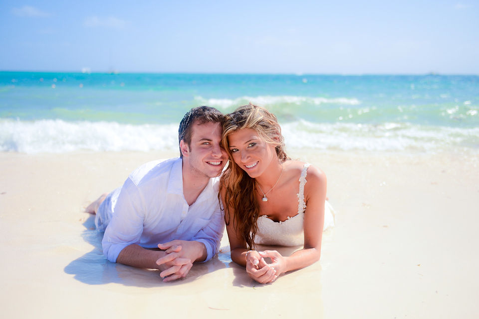 Pareja de recién casados acostados boca abajo, cabeza con cabeza y sonriendo frente al mar Caribe durante una sesión trash the dress en Punta Cana