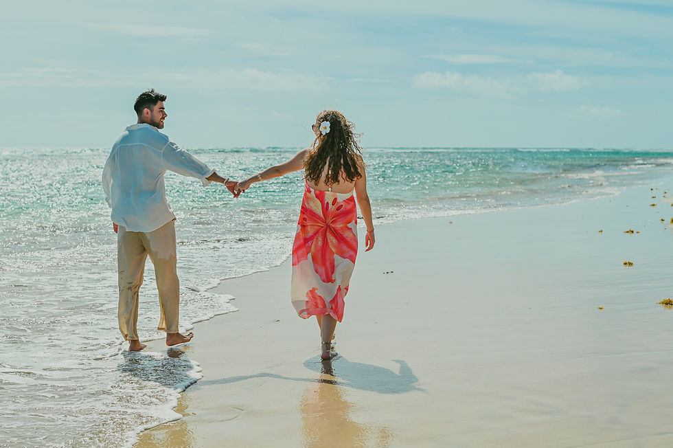Diego and Javiera walking away hand in hand along the shoreline of Uvero Alto Beach in Punta Cana Dominican Republic during a romantic couple photoshoot after their surprise marriage proposal, with the Caribbean Sea, blue skies and Javiera’s floral dress adding to this special engagement moment