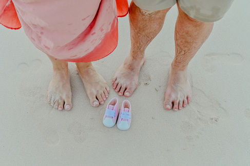 Foto de los pies de la pareja junto a los zapatitos de bebé en la arena durante una sesión de embarazo en Punta Cana