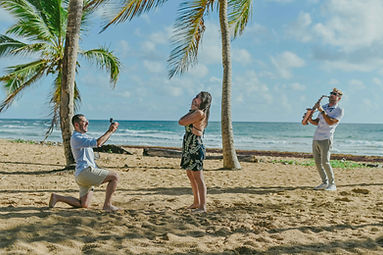 Hombre arrodillado mostrando un anillo de compromiso durante una propuesta de matrimonio sorpresa en Playa Uvero Alto Punta Cana mientras la futura novia se cubre la boca emocionada y un saxofonista toca junto al mar Caribe en Republica Dominicana