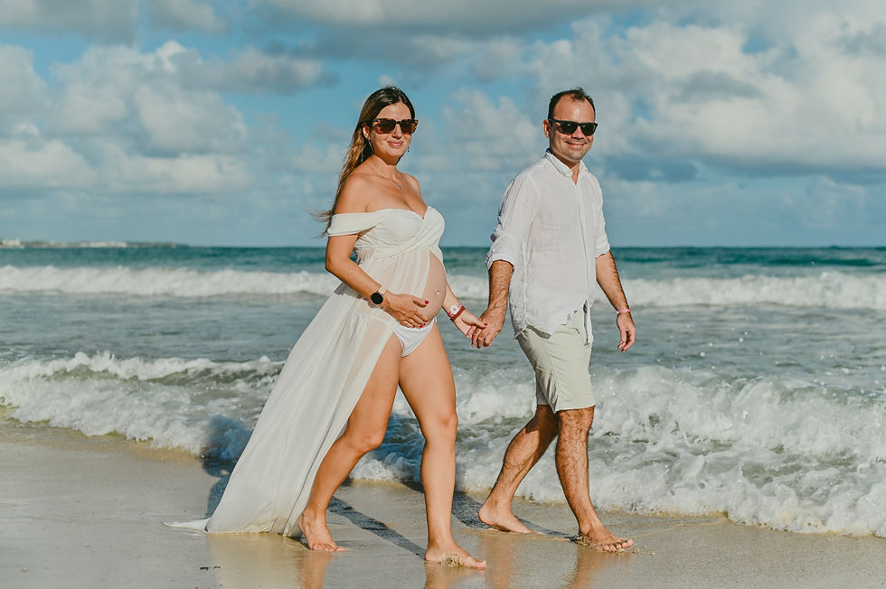 Pareja sonriente caminando en la playa durante sesion de fotos de embarazo en Punta Cana con mujer en vestido blanco bajo cielo azul con nubes en una atmosfera feliz y soleada