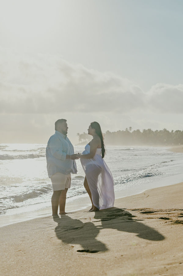 Backlit couple holding hands on the beach during a maternity photoshoot in Uvero Alto Punta Cana at sunset with white dress and romantic Caribbean atmosphere