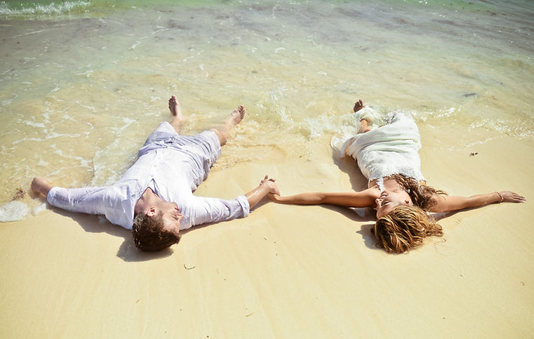 Pareja de novios recostada en la arena extendiendo las manos durante una sesion trash the dress en la playa en Punta Cana Republica Dominicana
