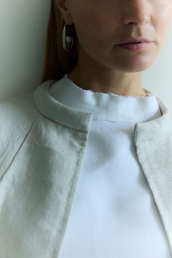 Close up of model wearing Chalk white duchess sweatshirt under Paperwhite silk cotton coat. She is wearing silver and pearl earrings and the multistitching detail around the neckline can be seen.