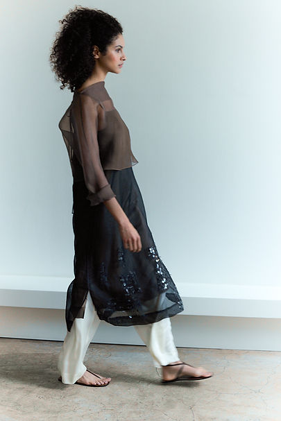 Third of set of three photos of model walking to the right in the atelier, wearing a charcoal organza sequinned slip dress with a nut brown organza T-shirt over the top and cream slipper satin PJ pants underneath. She is also wearing fine strappy sandals.