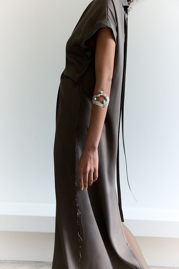 Cropped photo of side of model wearing a cocoa sueded satin pleat front shift dress with long back ties and a silver and pearl bracelet.