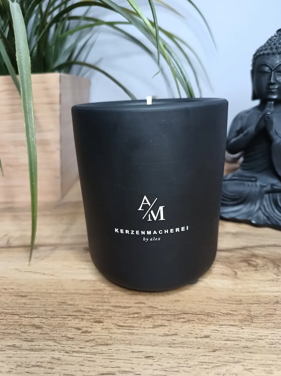 Miniaturbild: Schwarze Keramikkerze mit weißem Logo, AM, Keramikkerze Schwarz, Dekovision und Buddha-Statue.