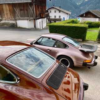 side rear view of two Porsche 911 G-Model, one with tail, old buildings and mountains in background