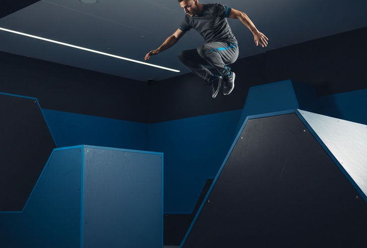A dynamic shot of a traceur performing a precision jump in an indoor parkour facility with blue and black obstacles.