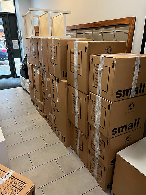 Large stack of cardboard moving boxes, wardrobe boxes, and small cartons organized along a tiled hallway corridor.