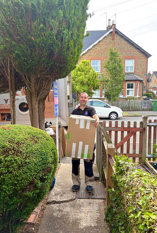 Experienced house removals operative carrying a sealed cardboard moving box down a garden path toward a transit vehicle parked outside a Victorian brick family home.
