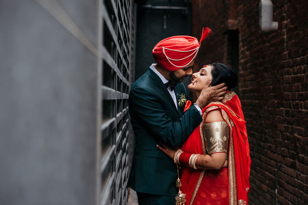 Photo by Monika Mistry Photography of a bride in a red outfit leaning against a textured wall in an urban setting.