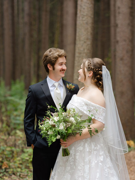 Bride and groom standing together smiling in forest with polished bridal hair and makeup.
