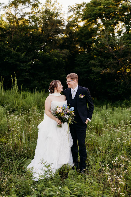 Bride and groom embracing in tall grass, bouquet, soft glam makeup and low updo.