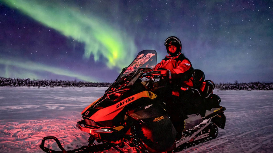 Evening snowmobile safari for families — children safely in a guide-pulled sled, kota hut stop on a frozen swamp, Arctic night sky above.