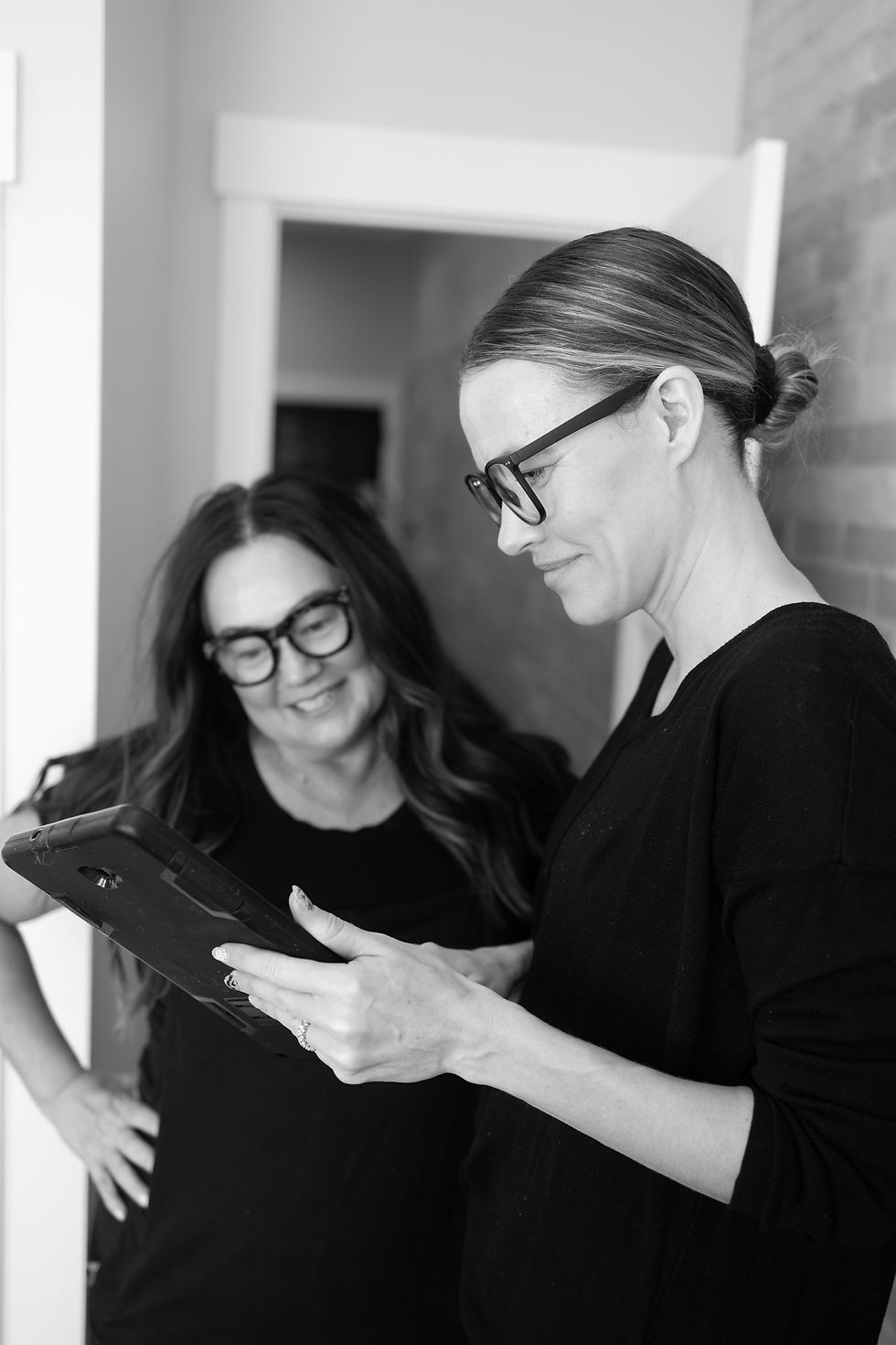 Two women wearing glasses look at a tablet, smiling. They're indoors, with a partially open door and brick wall in the background. Black and white.