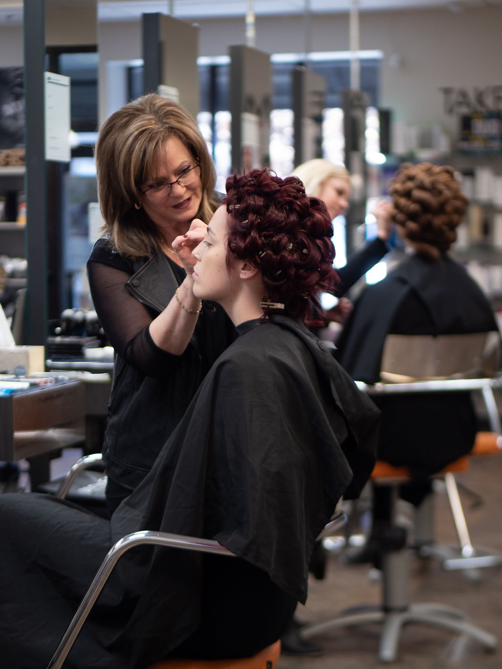 Stylist applies makeup to seated woman with curled hair in salon. Background shows another client and shelves. Calm, focused mood.